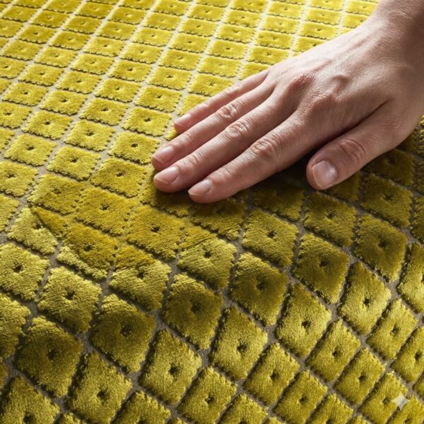 Eine Nahaufnahme einer Hand, die sanft über einen strukturierten, senfgelben Velours-Möbelstoff streicht. Das Muster besteht aus kleinen, erhabenen Quadraten mit einem punktförmigen Zentrum.